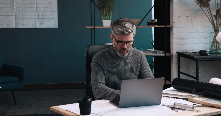 Serious and thoughtful Man working at his Office Room and typing on the Laptops Keyboard. Having tube with Drawings on the Desk. Middle-aged Man with Eyeglasses and stylish hairstyle. Royalty-Free Stock Footage #1059571199
