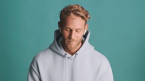 Attractive cheerful blond bearded man in hoodie and wireless earphones rejoicing and happily showing yes gesture over colorful background