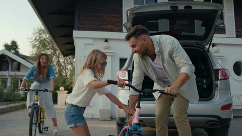Happy family spends time together on holidays. Attractive mom and pretty daughter having fun outside and riding bikes. Smiling dad in cottage garden got bicycle out of car for leisure of excited girl