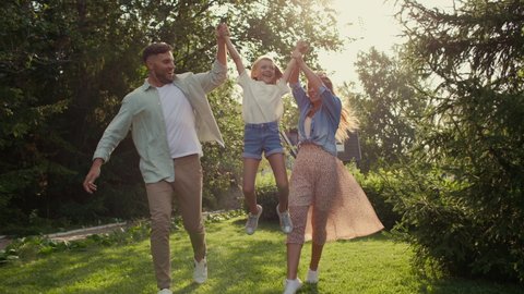 Happy playful family spends carefree time together outside in sun. Smiling dad and attractive mom with pretty daughter having fun and walking near house. Leisure of excited girl at holidays in nature