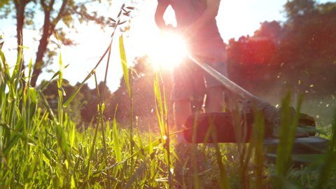 SLOW MOTION, MACRO, LOW ANGLE, LENS FLARE, DOF: Guy uses a handheld grass trimmer to landscape his garden on a sunny spring day. Unrecognizable man uses a petrol scythe to cut the grass in his yard.