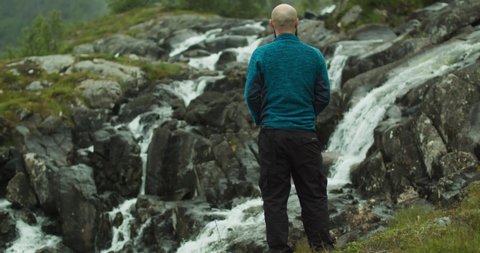 A traveler in a blue sweatshirt stands by a beautiful Norwegian waterfall and observes the wonderful nature of the fjords. Slow motion footage.