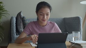 Happy Asian woman waving or greeting online tutor on tablet in the living room at home. Concept online learning at home