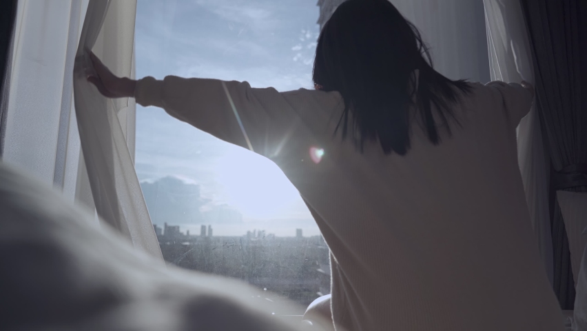 Silhouette Shot from behind of Young Asian woman open window curtain to enjoy beautiful day light, sitting down on bed with beautiful morning sunshine, deep sleep getting up late on weekends