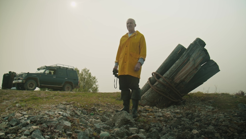 An elderly man with gray hair in a yellow raincoat near an SUV, looking through binoculars at sunset in smog. An adult man is looking for new places to travel by car. Active ageing. Slow motion, 4K
