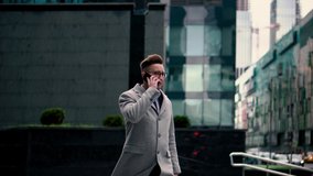 A fashionable young man in a coat and glasses is walking in the center of the city among skyscrapers and talking on the phone
