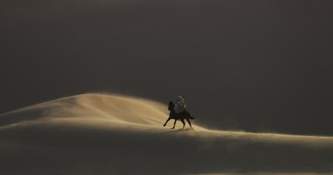 Masked person rides a galloping horse through the desert in a sand storm. Sand dunes surround a person riding a horse with a rifle on their back. Long journey. Cinematic, Shot on RED