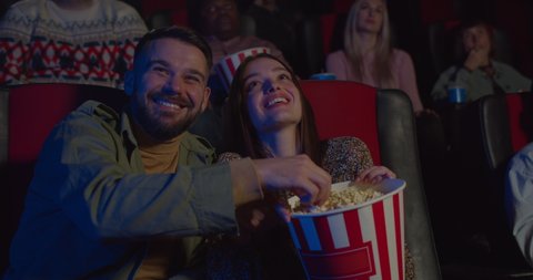Close up of girlfriend stealing popcorn from her boyfriend hand while watching movie in cinema.Couple having good time and laughing while having date in cinema