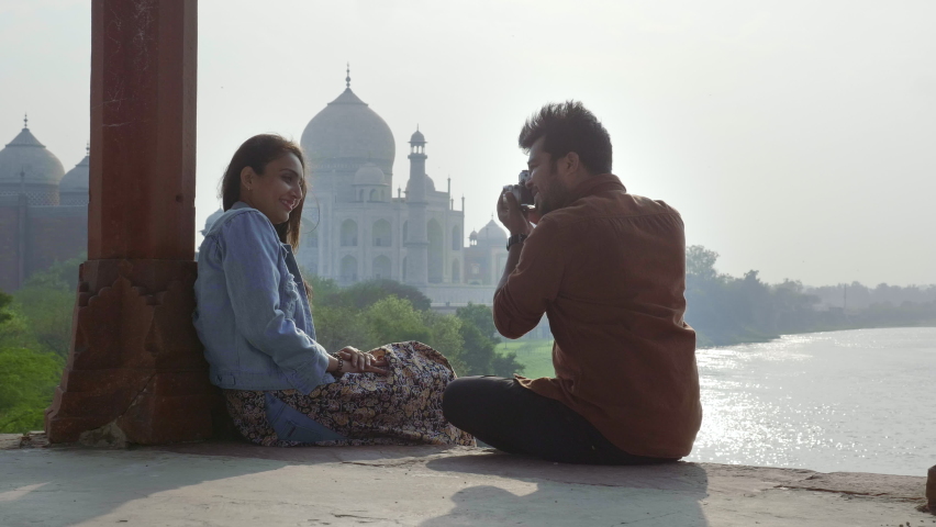 A happy and young romantic couple is sitting outdoors against Taj mahal and girlfriend is posing for boyfriend who is clicking pictures using a camera. Cheerful male and female enjoying time together