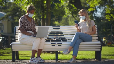 Young diverse women keeping social distancing sitting on bench in park using gadgets. Multiethnic college students sitting on campus bench in safety mask surfing internet on laptop and smartphone