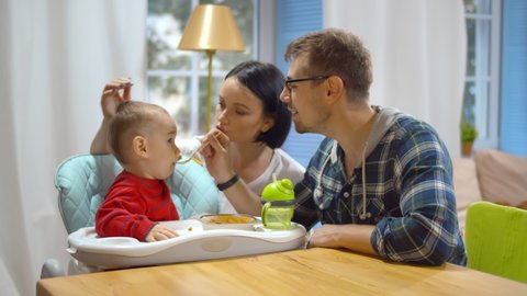 Happy parents feeding baby son with healthy porridge sitting at kitchen. Happy smiling mother and father giving food to cute little boy in highchair. Family and parenthood concept