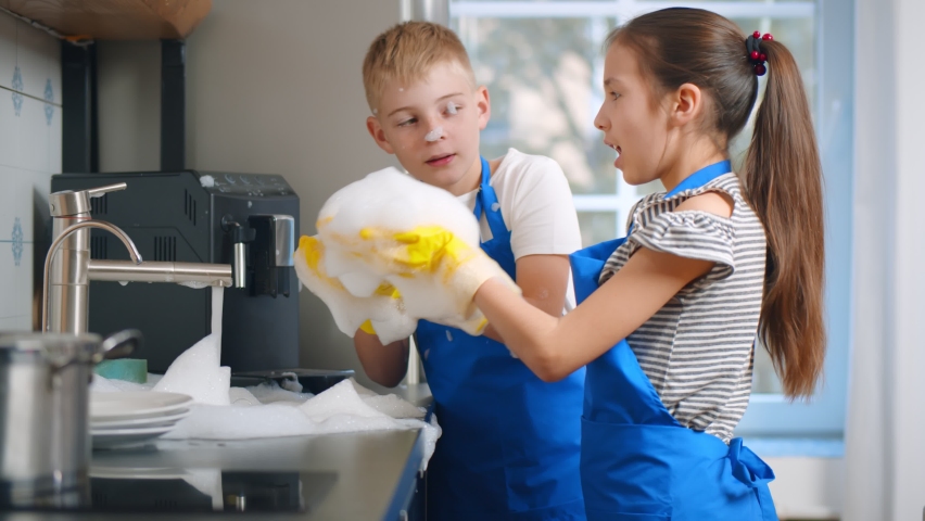 Happy boy and girl kids washing dishes and playing with foam in kitchen. Brother and sister children in apron and gloves cleaning plates and having fun blowing soap foam Royalty-Free Stock Footage #1059597065