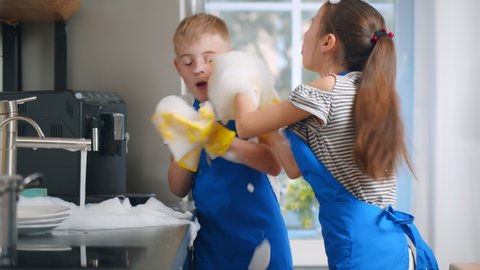 Happy boy and girl kids washing dishes and playing with foam in kitchen. Brother and sister children in apron and gloves cleaning plates and having fun blowing soap foam