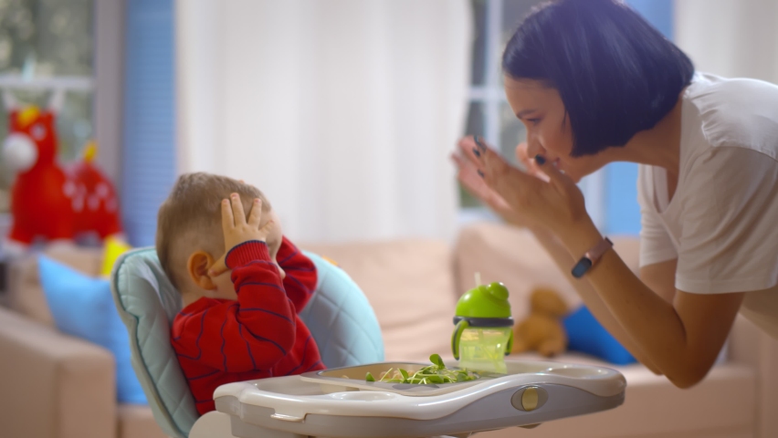 Mother and baby boy playing peekaboo and have fun at home. Side view of happy woman playing peek-a-boo with toddler boy sitting on highchair. Motherhood and family concept Royalty-Free Stock Footage #1059597074