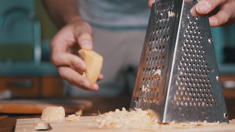 Man Hand Rubs Hard Cheese on a Metal Grater. Cheese Crumbles Smoothly onto a Cutting Board. Home kitchen. Close-up. Slow Motion.