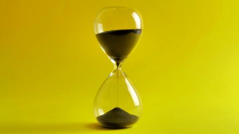 Shot of a sand clock measuring time while the sand is falling down against the yellow background - old classic timer. Extreme close up of a transparent hourglass with flowing black sand - time concept
