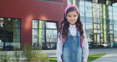 Portrait of cute beautiful Caucasian little school girl smiling while standing in front of school. Pretty nice child in good mood outdoors. Adorable female junior student. Back to school concept