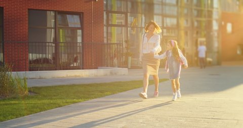 Beautiful Caucasian cheerful mother walking with cute small schoolgirl outdoors on sunny day from school and having fun. Happy woman with little kid running and jumping on street in city. Fun concept
