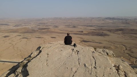 Zooming out on a guy sitting on crater's edge in the middle of the desert at the Negev, Israel