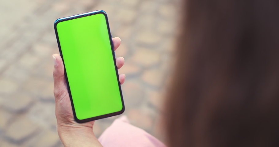 Over shoulder view of female person holding and pressing on smartphone with mockup screen while sitting at street. Concept of chroma key and greenscreen Royalty-Free Stock Footage #1059623042