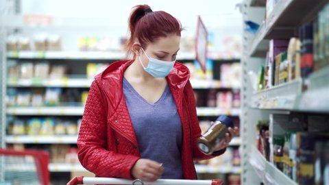 Red haired woman in red jacket wearing medical mask choosing jar of instant coffee at grocery store and putting it in shopping cart, blurred people passing by on background. Concept of pandemic