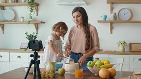 A Pretty Mother With a Cute Daughter Shooting a Blog About Cooking. Video Blog About Healthy Food. A Mother With Child Streaming Online Video Vlog. Lovely Mother Teaching Her Daughter to Cook.