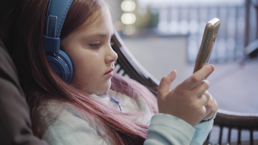 Absorbed concentrated Caucasian girl sitting in hanging chair with smartphone and surfing Internet. Close-up portrait of cute device addicted kid in headphones messaging online.