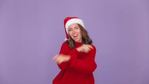 Jumping woman 20s in red sweater Santa Christmas hat dancing fooling around having fun expressive gesticulating hands isolated on purple violet background studio. Happy New Year merry holiday concept
