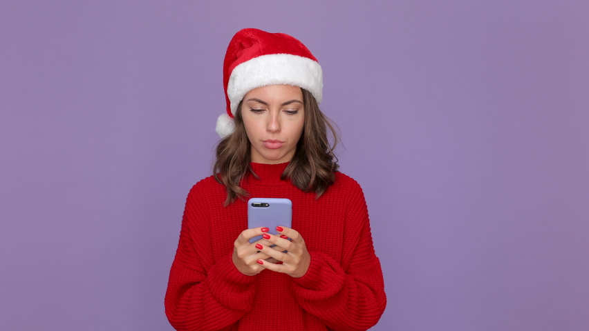 Shocked woman in red knitted cozy sweater Santa Christmas hat holding using mobile cell phone typing sms isolated on purple violet background studio. Happy New Year celebration merry holiday concept | Shutterstock HD Video #1059642935