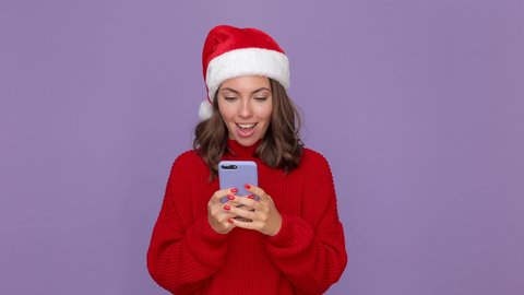 Shocked woman in red knitted cozy sweater Santa Christmas hat holding using mobile cell phone typing sms isolated on purple violet background studio. Happy New Year celebration merry holiday concept