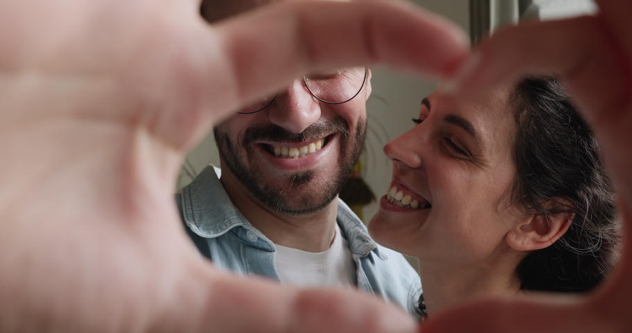 Close up portrait happy couple faces looking through joined fingers making heart shape. St Valentines Day celebration, romantic relationships, just married spouses, sincere feelings and love concept | Shutterstock HD Video #1059643868