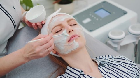 Medium shot of Beautician putting cream mask on woman's face at beauty salon. Facial skin care treatments.