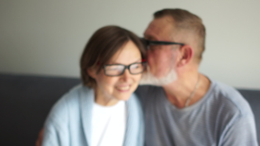 Happy elderly couple in love. Old man whispering secret to his wife. Gray-haired man and woman are sitting indoors on the sofa. Woman wears glasses and laughs cheerfully Royalty-Free Stock Footage #1059646370