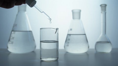 Close-up of a pipette in hand and a glass beaker against the background of flasks in the laboratory. A colorless liquid drips into a glass. Perfumery production.