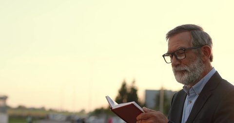 Old wise Caucasian gray-haired man in glasses walking and reading book outdoors in sunset at street. Senior clever male professor in eyeglasses strolling and read textbook. Studying by walk.