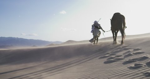 Masked person leads a horse through a sand storm in the desert. Person with a rifle on their back walking. Cinematic, Shot on RED