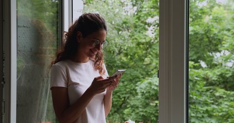 Attractive young woman in glasses standing near open window holding smart phone looking outside enjoy view admires warm summer day lush green trees, using internet communication chat feels carefree