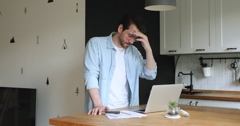 Man standing in home kitchen use calculator calculates personal finances looks at laptop screen app feels stressed and anxious about bank overspend, mistake in financial report, bankruptcy concept