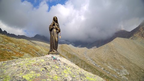 Val Masino, Province of Sondrio Italy, September 2020: Bronze statue of Mary Magdalene atop an alpine slope in the Italian Alps. Madonna with a cross in her hands. White clouds over the mountains