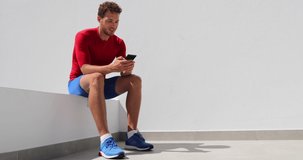 Fitness Athlete man texting looking at phone screen at home after working out training. Technology and sports athlete holding mobile phone. Happy fit young adult in compression clothing sportswear.