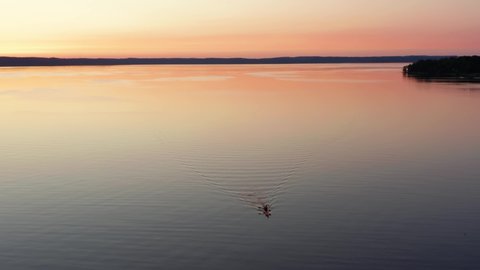 Sunset kayaking on a lake. Aerial drone view of young man kayaker on water with yellow orange purple pink night light at horizon. Sport activity out in the wild nature on calm summer evening. Sweden