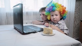 Little caucasian children in cone hat and funny wig sit at table with laptop computer, talk to relatives via video call. father lights candle on birthday cake. party at home during covid-19 quarantine