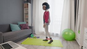 Young african american woman hippy in colourful sportswear with curly afro hairs does exercise on legs with elastic band and watches fitness video tutorial online on laptop computer