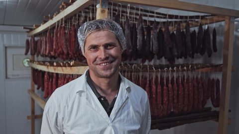 Portrait of professional butcher meat specialist standing inside smokehouse, smiling cheerful looking tired after working shift. Meat processing factory. People.