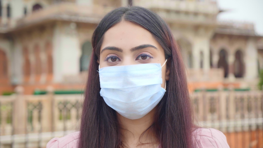 A close up shot of a young beautiful woman or female standing outdoors wearing a protective face mask looking at the camera amid Corona Virus COVID 19 epidemic or pandemic Royalty-Free Stock Footage #1059671783