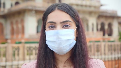 A close up shot of a young beautiful woman or female standing outdoors wearing a protective face mask looking at the camera amid Corona Virus COVID 19 epidemic or pandemic