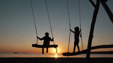 Silhouettes of Two Children, Little Friends Swaying on Rope Swings Near Sea at Sunset. Calm Relax Together, Summer Happiness, Dream Freedom of Girl Enjoying Carefree Childhood, Having Fun Boy Outdoors