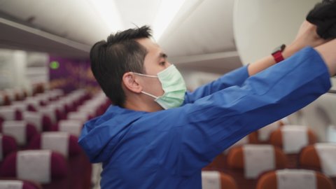 Asian male traveler wearing surgical mask walks in airplane aisle finding seat in passenger plane cabin during covid pandemic. Man carrying bag and put in the overhead storage then sit on window seat.