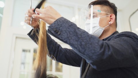 Professional male stylist cutting woman's hair in salon. The man wearing mask and face shield to prevent from coronavirus infection during pandemic. Beauty salon business concept.
