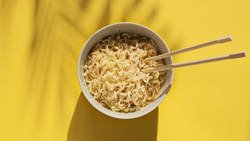 Instant noodles are mixed with chinese chopsticks on yellow background. Female hand mixes a hot meal. Top view egg noodles with spices in plate bowl. | Shutterstock HD Video #1059680249